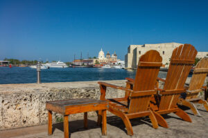 Panoramic of the Historic Center of Cartagena with wooden chairs in front of the La Bodeguita pier, the imposing colonial walls and the San Pedro Claver cathedral in the background, next to the Convention Center on the right, capturing the historical and cultural essence of this Caribbean city.