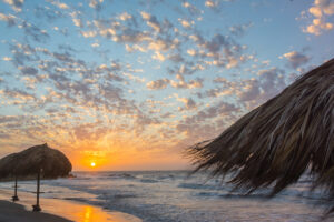 A beach scene at sunset with thatched-roof umbrellas and a sky filled with scattered clouds illuminated by the setting sun.